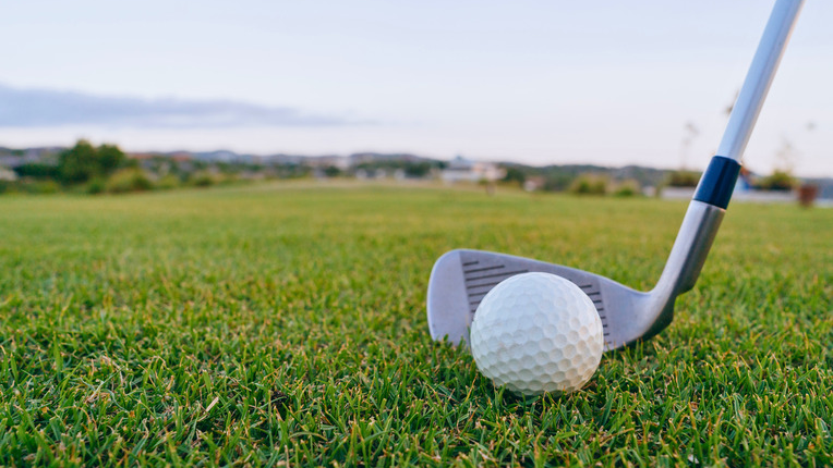 A golf ball on a lush green golf course with a gold club in the background.