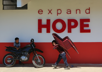 the words expand hope on a building with a boy leaning on motorcycle and another boy with plastic chairs on his back